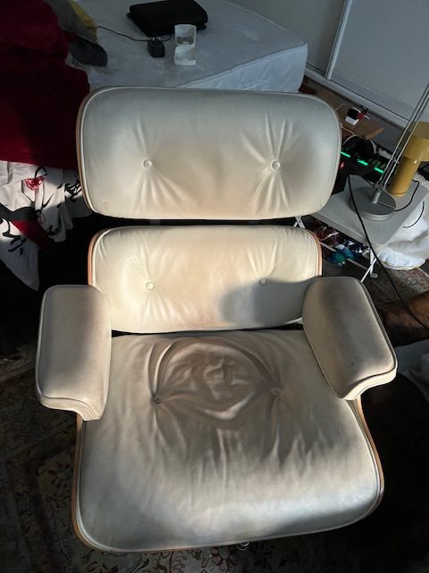 A white upholstered lounge chair with a high backrest and armrests, showing signs of wear with visible creases and soft cushions, positioned in a cluttered room.